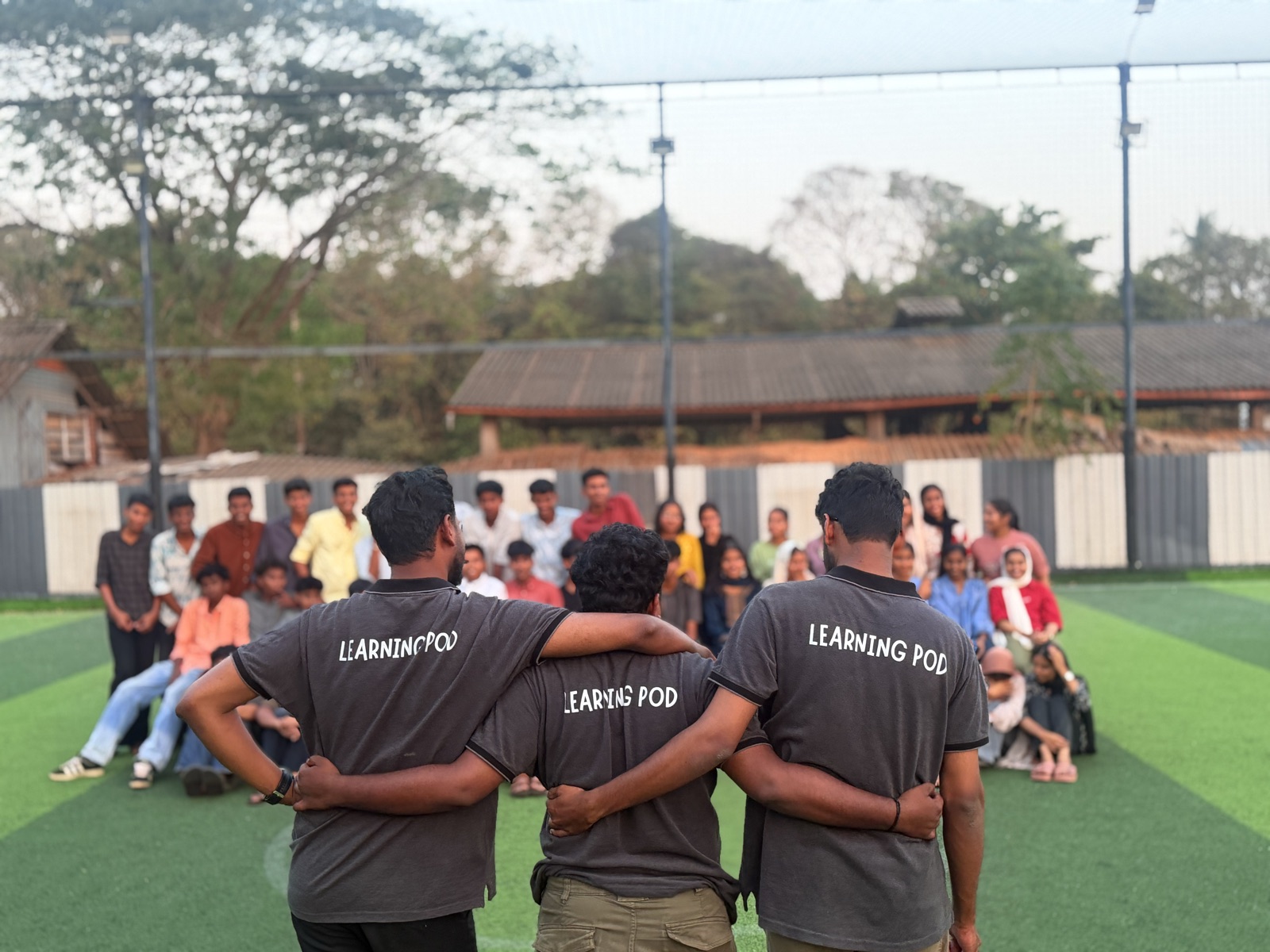 Three Learning Pod tutors arm-in-arm facing their seated students on the turf